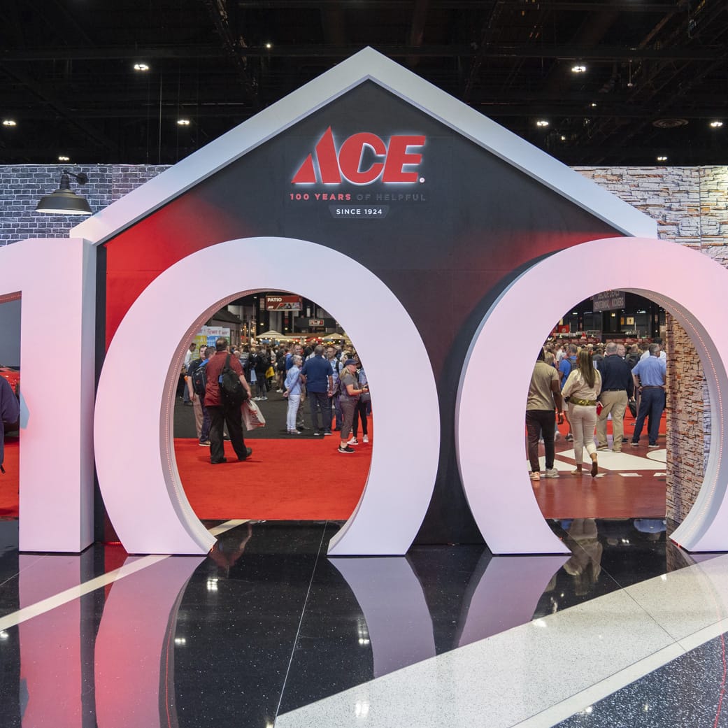 Large ACE Hardware display shaped like a house with “100” in big white numbers, celebrating 100 years. People can be seen walking through and gathering inside the event space.
