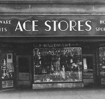 Black and white photo of an old storefront with a large sign reading "ACE STORES." The window displays various hardware items, and smaller signs are partially visible on both sides of the entrance.