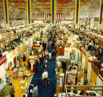 A large, crowded indoor trade show or convention with many booths, displays, and people walking through wide aisles under a high ceiling with yellow and blue banners.