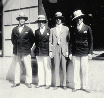 Four men stand side by side on a city sidewalk, all wearing straw boater hats, dark jackets, light trousers, and ties, with one in a lighter suit, in a vintage black and white photograph.