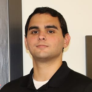 A young man with short dark hair, wearing a black collared shirt over a white undershirt, stands indoors against a pale wall, looking directly at the camera with a neutral expression.