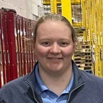 A person with light skin and blonde hair tied back is smiling, wearing a blue collared shirt and dark jacket, standing in a warehouse with red lockers and yellow shelving in the background.