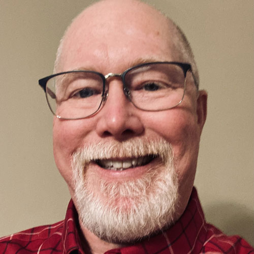 A smiling older man with glasses, a white beard, and a bald head, wearing a red plaid shirt, posing in front of a plain beige background.