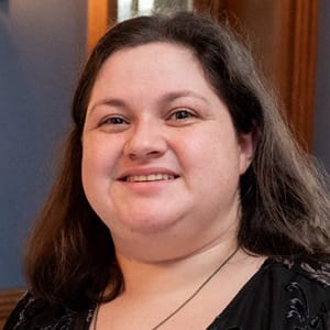 A woman with long brown hair smiles at the camera. She is wearing a black top with a subtle pattern and a necklace. The background shows a wooden wall and soft indoor lighting.