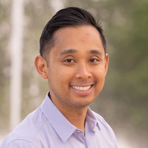 A man with short dark hair, wearing a light purple collared shirt, smiles at the camera. The background is blurred with greenery and light colors.