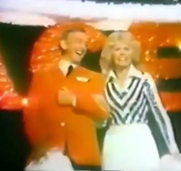 A man in a bright orange jacket and a woman in a black-and-white patterned top stand smiling together on a brightly lit stage with large, illuminated orange letters in the background.