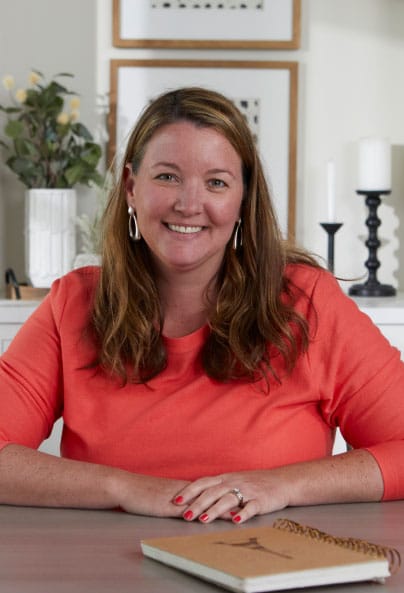 A woman with long brown hair, wearing a bright orange top, sits at a table with a notebook in front of her. She is smiling, with her hands folded, in a room decorated with candles, artwork, and a vase of flowers.