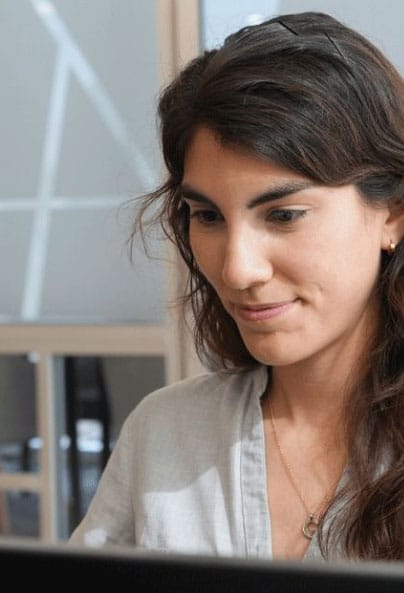 A woman with long dark hair and a light-colored shirt is sitting indoors, looking at a computer screen and smiling slightly. She appears focused and engaged in her work.