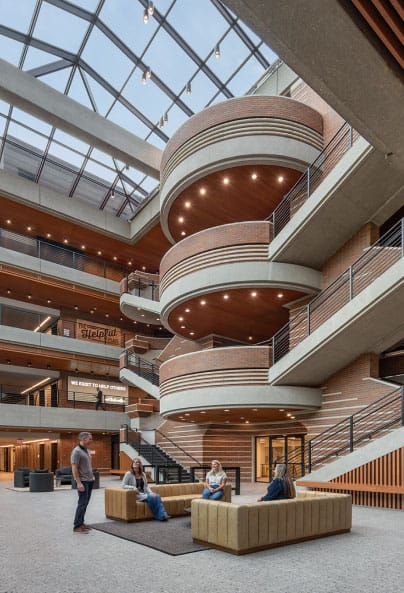 Four people converse while seated on modern couches in a spacious, multi-story atrium with large circular balconies, exposed brick, glass ceiling, and abundant natural light.
