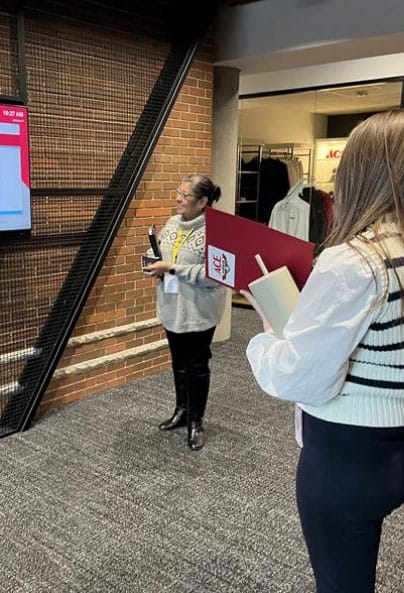 Two women stand indoors near a brick wall; one holds a notebook and pen, the other looks at a large screen with a presentation displayed. Clothing racks are visible in the background.