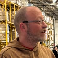 A man with glasses and a beard, wearing a brown hoodie, stands in a warehouse with shelves of boxes behind him.
