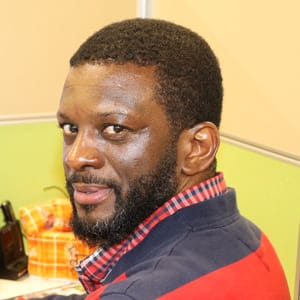 A man with short black hair and a beard, wearing a red and blue plaid shirt, sits at a desk in an office cubicle, looking over his shoulder and smiling slightly at the camera.