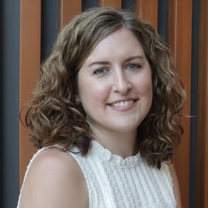 A woman with curly, light brown hair and a white sleeveless top smiles at the camera. She stands in front of a background with vertical wooden and dark panels.