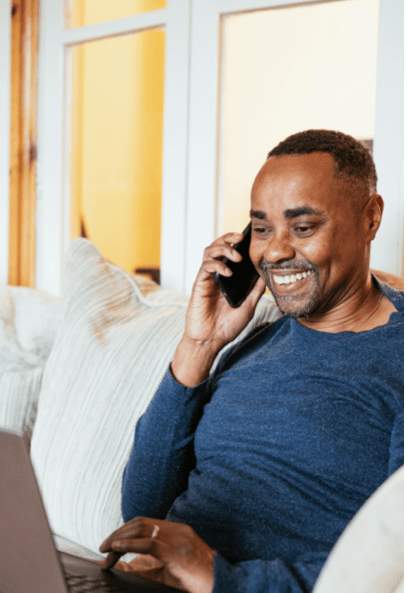 A man sitting on a couch, smiling while talking on a smartphone and using a laptop.
