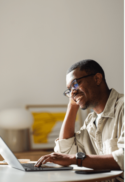 A smiling man wearing glasses and a beige shirt sits at a table, working on a laptop. He rests his head on one hand and appears relaxed. A framed abstract artwork and lamp are blurred in the background.