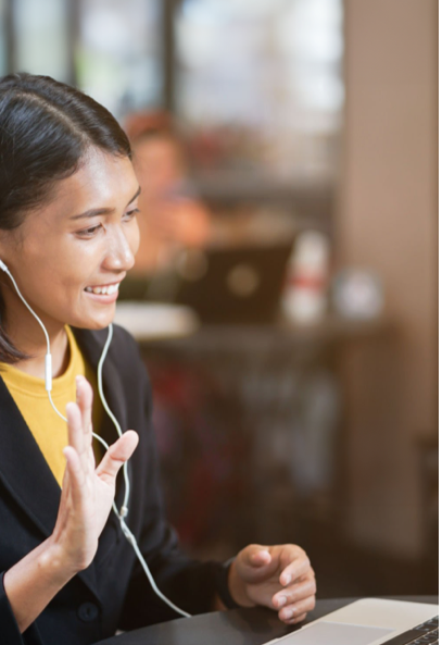 A woman with earphones is smiling and waving while video chatting on a laptop in a cozy indoor setting. The background is blurred, suggesting a cafe or shared workspace.