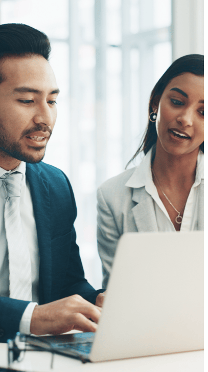 Two people in business attire sit together at a desk, looking at a laptop screen and discussing something. The background shows large windows with natural light.
