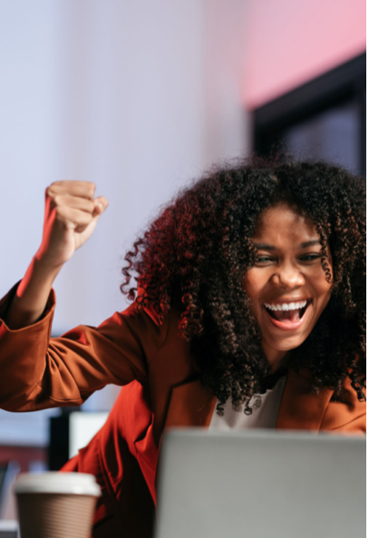 A woman with curly hair smiles and raises her fist in excitement while sitting at a desk with a laptop and a coffee cup in front of her.