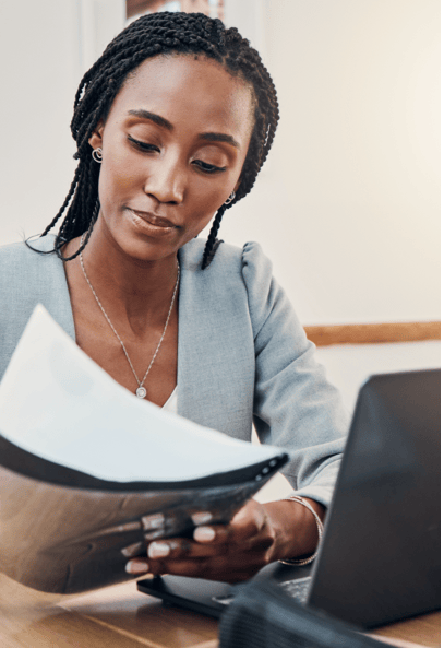 A woman with braided hair, wearing a light gray blazer, sits at a desk looking through papers next to an open laptop.