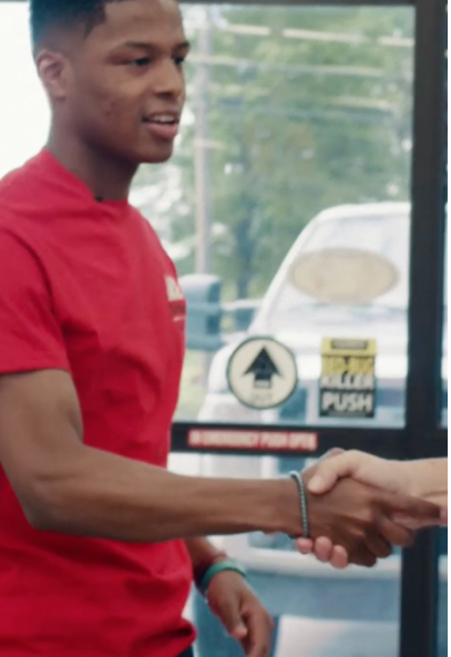 A young man in a red shirt shakes hands with someone in front of a glass door with signs, including one that says "PULL" and another that says "PUSH." A car is visible outside through the glass.