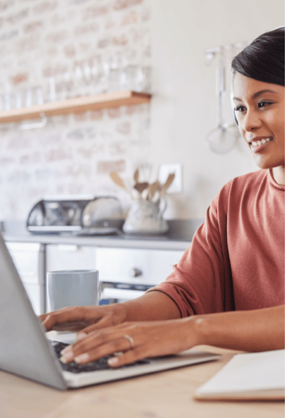 A woman sits at a kitchen table, smiling and typing on a laptop. A coffee mug is next to her, and shelves with kitchen items are visible in the background.