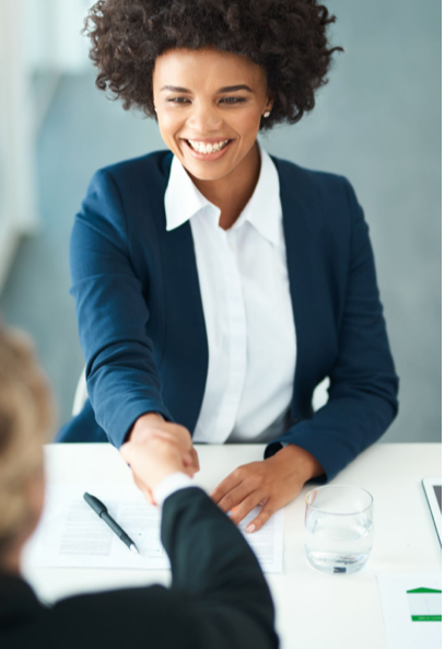 A smiling woman in a blue blazer and white shirt sits at a desk, shaking hands with another person across the table; paperwork and a glass of water are on the desk.