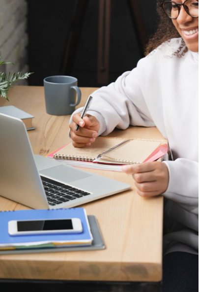 A person smiles while sitting at a desk with a laptop, notebook, pen, and smartphone. They appear to be studying or working, with a coffee mug and books nearby.