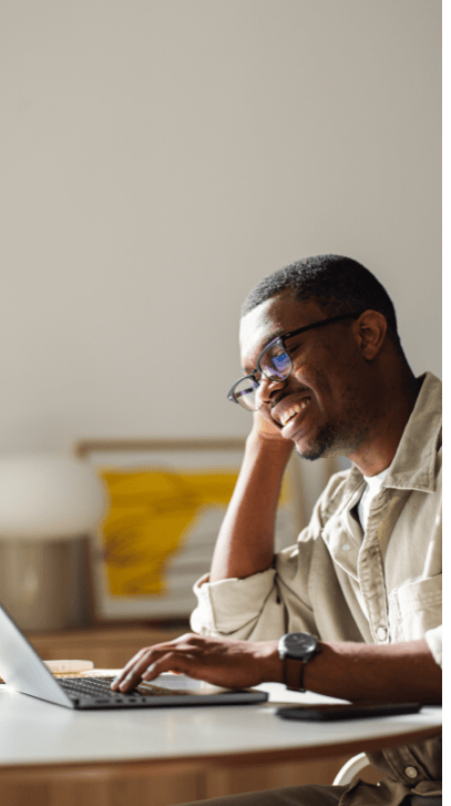 A smiling man wearing glasses and a beige shirt sits at a desk, resting his head on his hand while working on a laptop. A framed abstract artwork and lamp are blurred in the background.