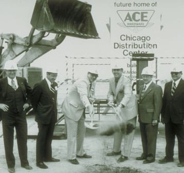 Six men in suits and hard hats take part in a groundbreaking ceremony, shoveling dirt in front of a sign that reads "Future Home of ACE Hardware Chicago Distribution Center" with construction equipment behind them.
