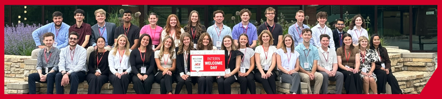 A large group of interns sits outside on a stone bench, smiling at the camera. Several hold a sign reading "Intern Welcome Day." The group is dressed in business casual attire, with greenery and a building behind them.