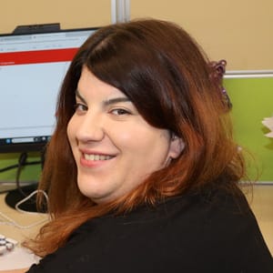A woman with long brown hair is smiling at the camera while sitting at a desk with a computer monitor and office supplies in the background.