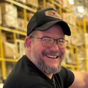 A smiling man with glasses, a beard, and a black cap stands in front of yellow warehouse shelves filled with boxes.
