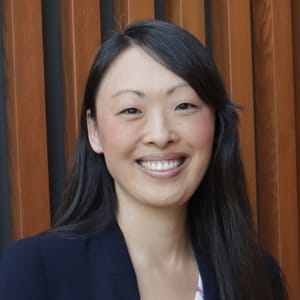 A woman with long dark hair smiles at the camera, standing in front of vertical wooden slats. She is wearing a dark blazer and a light-colored top.