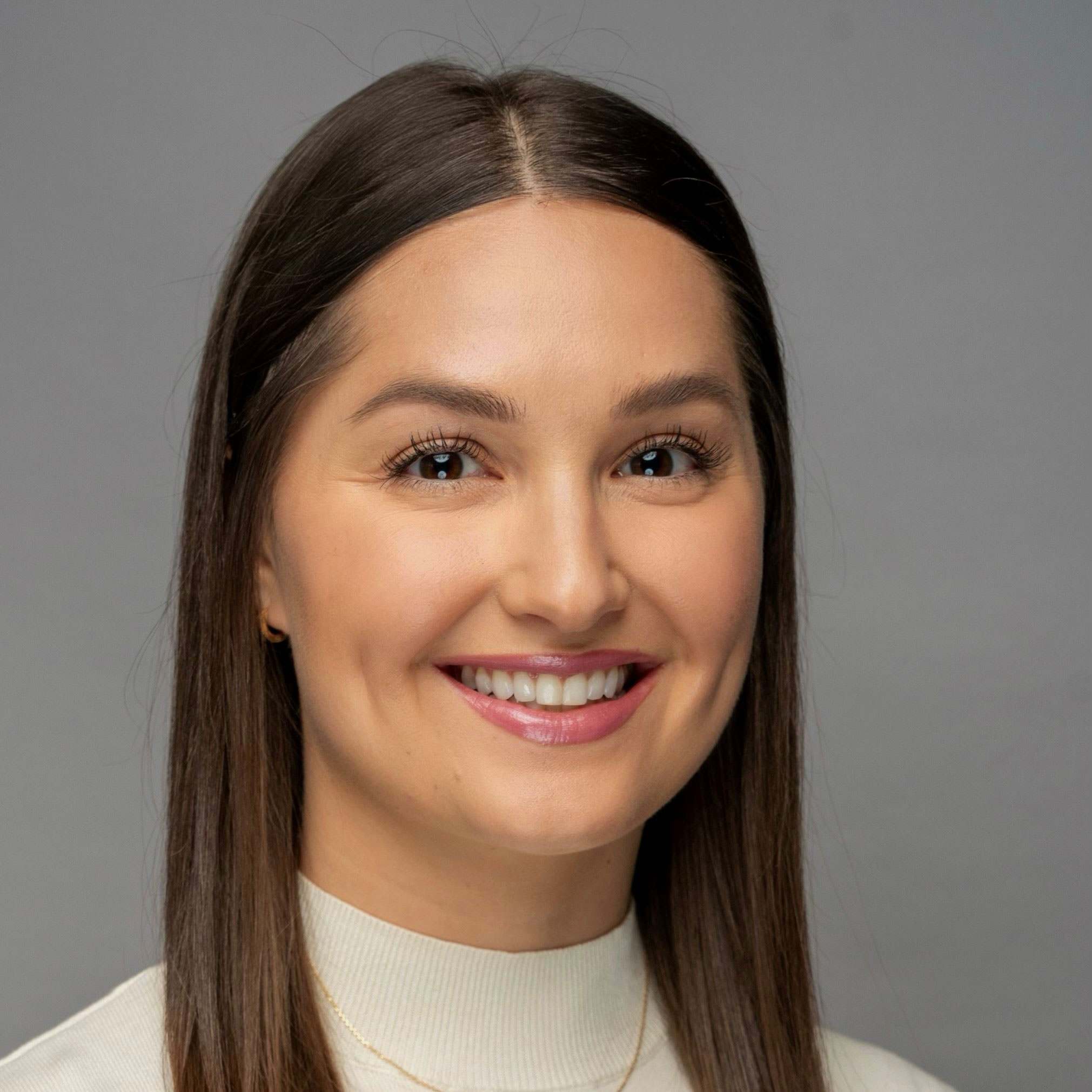 A woman with straight brown hair, wearing a light-colored turtleneck sweater and a thin necklace, smiles at the camera against a plain gray background.