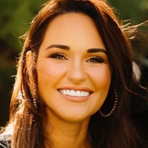 A woman with long brown hair and hoop earrings smiles brightly outdoors, with a blurred green background behind her.