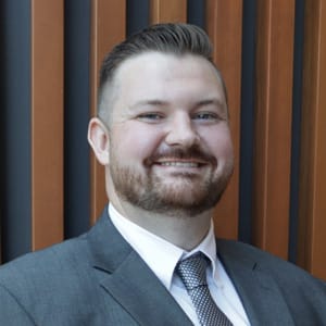A man with short brown hair and a beard, dressed in a gray suit, white shirt, and patterned tie, smiles at the camera. The background features vertical brown and black striped panels.