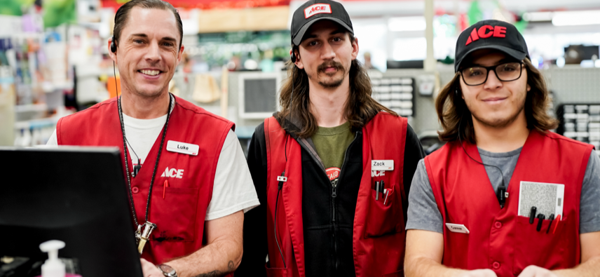 Three Ace Hardware employees wearing red vests and name tags stand together behind a store counter, smiling at the camera. The background shows shelves and store merchandise.