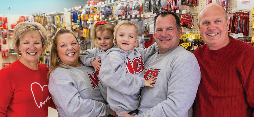 A smiling family of six stands in a hardware store aisle. Four wear matching gray "ACE" sweatshirts, while two wear red. Tools and hardware hang on the wall behind them. Two young children are held by adults.