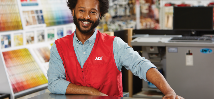 A smiling employee wearing a red ACE vest stands at a counter in a hardware store, with colorful paint samples and store equipment in the background.