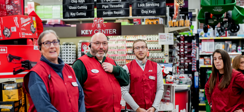 Four hardware store employees wearing red vests stand together and smile in front of the "Key Center" area, surrounded by shelves of merchandise and signs for services like key cutting and screen repair.