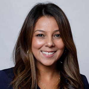 A woman with long brown hair and a friendly smile, wearing a dark top, poses in front of a plain light gray background.