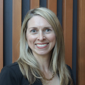 A woman with long blonde hair, wearing a black top and a gold necklace, smiles at the camera. She stands in front of a background with vertical wooden slats.