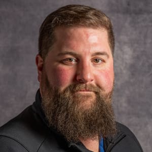 A man with short brown hair and a thick, full beard is wearing a dark jacket and smiling slightly while posing against a gray textured background.