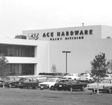 Black and white photo of an ACE Hardware Paint Division building with a parking lot in front, several parked cars, a few trees, and the ACE Hardware logo on the building.