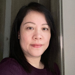A woman with straight black hair and light skin is standing indoors in front of a textured background, wearing a dark purple top and looking at the camera with a neutral expression.