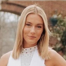 A young woman with straight, shoulder-length blonde hair smiles slightly while wearing a white, sleeveless lace top. The background is outdoors with blurred greenery and a brick structure.