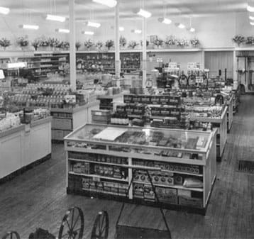 Black and white photo of a vintage grocery store interior with shelves and display cases filled with goods, wooden floors, and floral decorations lining the top of the back walls.
