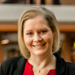 A woman with blonde hair, wearing a black blazer over a red top, smiles at the camera. The background is softly blurred, featuring warm indoor lighting.