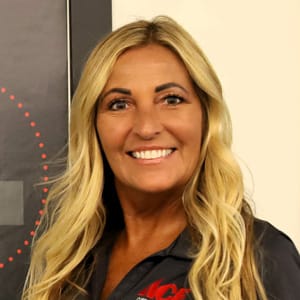A woman with long blonde hair smiles at the camera. She is wearing a dark collared shirt and standing in front of a light-colored wall and a framed picture with a dotted red circle.