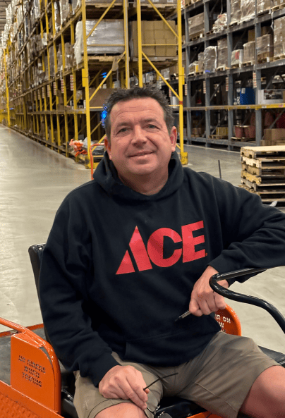 A man wearing a black hoodie with a large red "ACE" logo sits on an orange cart in a warehouse aisle lined with tall shelves full of boxes and pallets. He is smiling and holding eyeglasses.